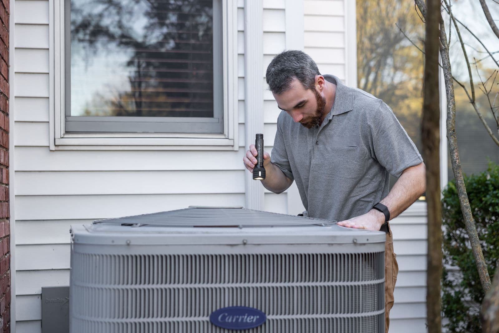 HVAC technician using a flashlight to inspect a Carrier residential central air conditioner condenser at the side of a home